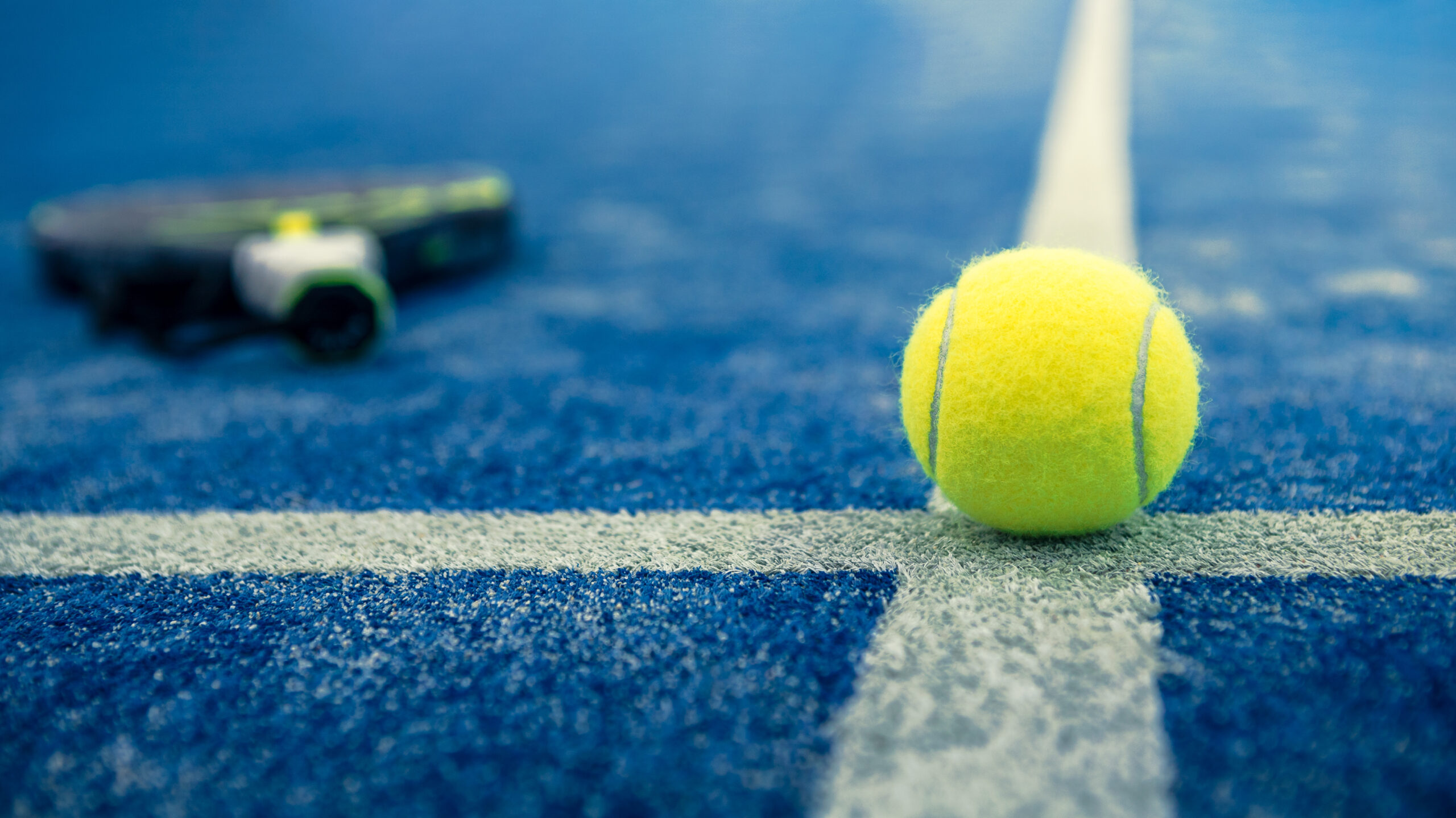 Yellow ball on floor behind paddle net in blue court outdoors. P